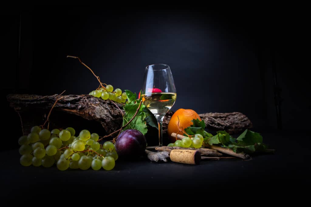 Vin Blanc et Fruits : Nature Morte Élégante et Rustique Verre de vin blanc, grappes de raisins verts, prune et orange sur fond noir rustique avec un bouchon de liège.