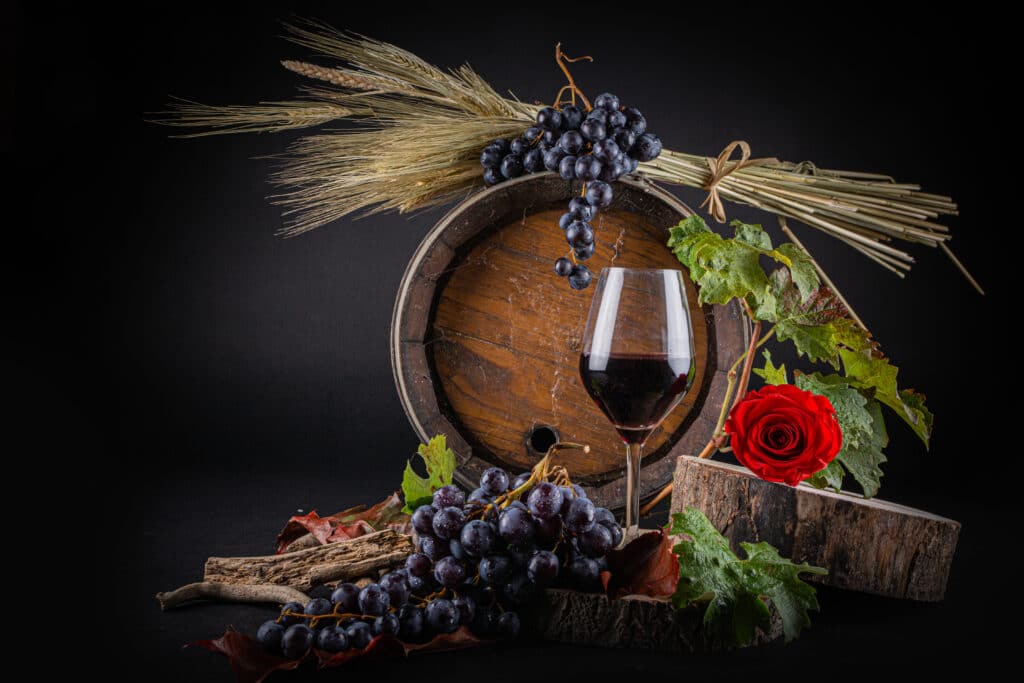 Vin Rouge, Raisins et Tonneau : Composition Rustique Nature morte avec verre de vin rouge, raisins noirs, tonneau en bois, blé et rose rouge sur fond noir.